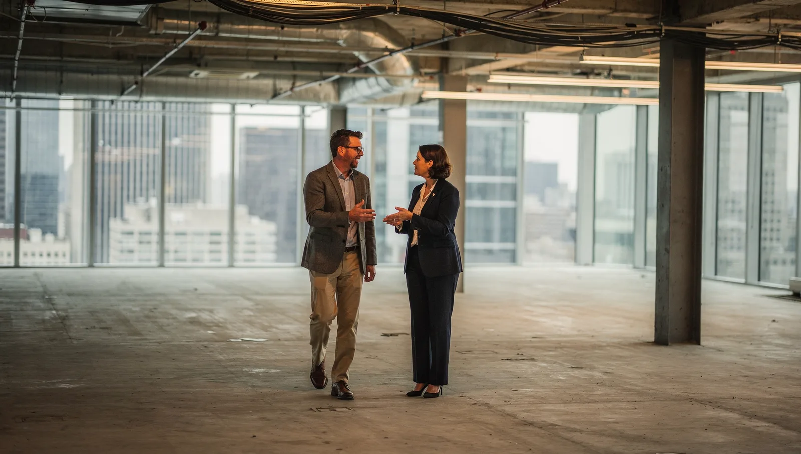 A man and woman in professional suits walk through an empty, raw commercial office space with skyline views.